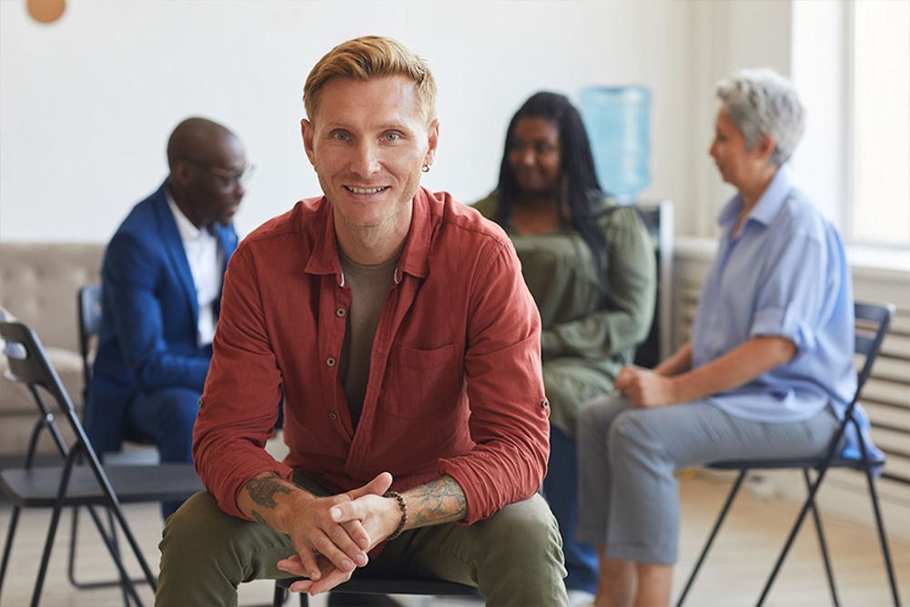 group-therapy-alcohol-addiction Smiling man looking out from mixed gender support group for alcohol addiction.